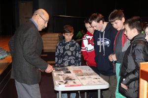 an older gentleman talks to a group of young boys as they surround a table filled with documents