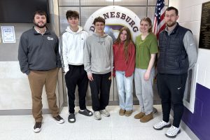 two male adults pose with four teenaged students in front of a school logo