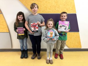 four elementary students pose holding books in the hallway of a school