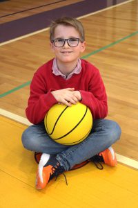 a boy wearing a red sweater sits on a gym floor and holds a yellow basketball