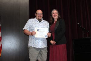 a bald man wearing a short sleeve shirt and shorts holding a certificate stands next to a woman wearing a red dress and black coat