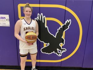 a girl holds a basketball in front of a purple wall with the letter D and an eagle, in a gymnasium