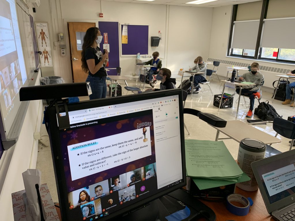 A computer screen in the forefront of view of a classroom with a teacher standing in front and students at desks