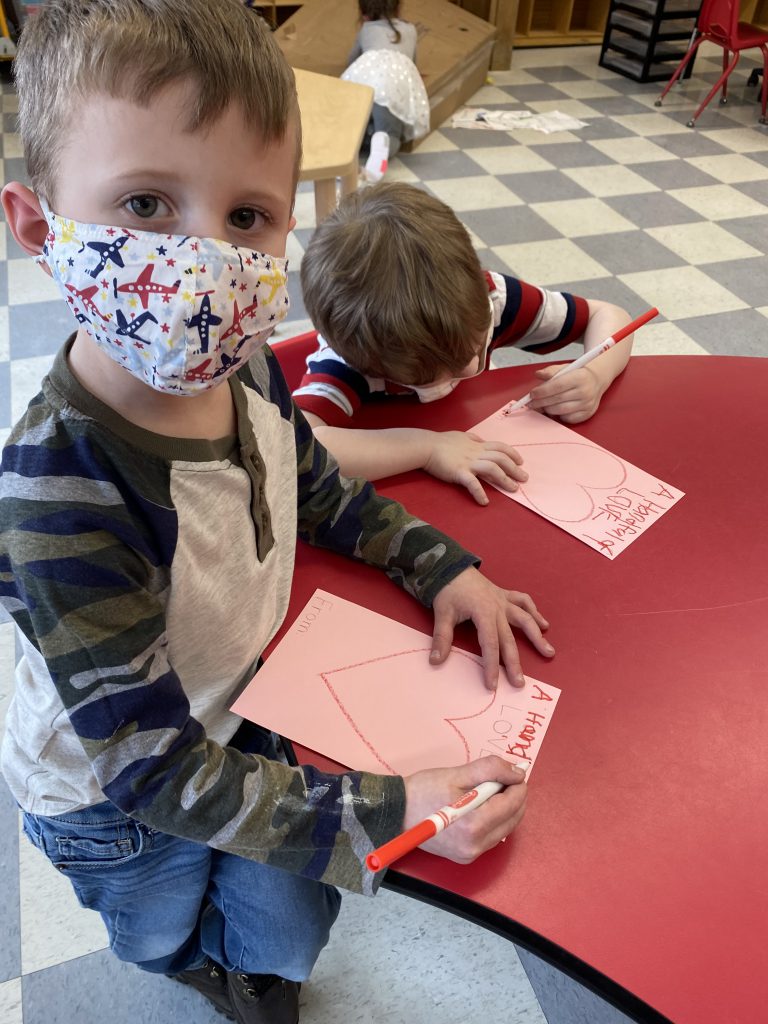elementary student wearing a mask uses a marker to create a valentine and stands next to a desk while a seated classmate works on their own valentine