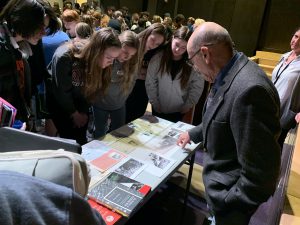 a group of teenage girls look at documents displayed on a table, as an older gentleman speaks