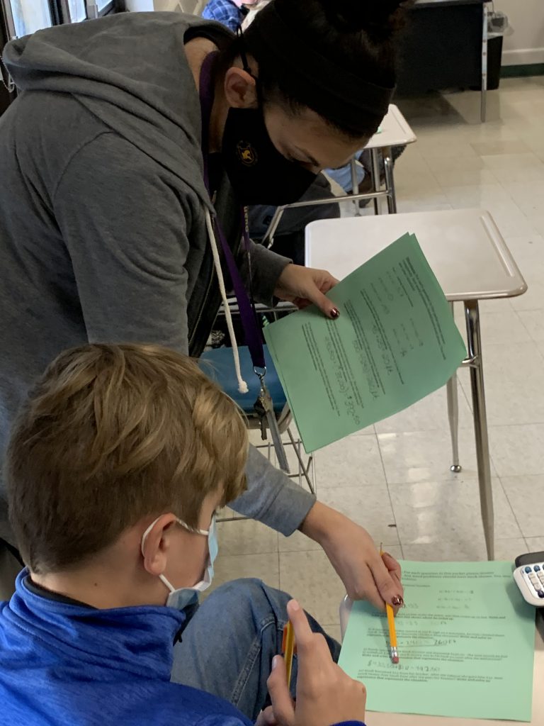 an educator wearing a mask standing next to a student's desk points to a piece of paper with a pencil