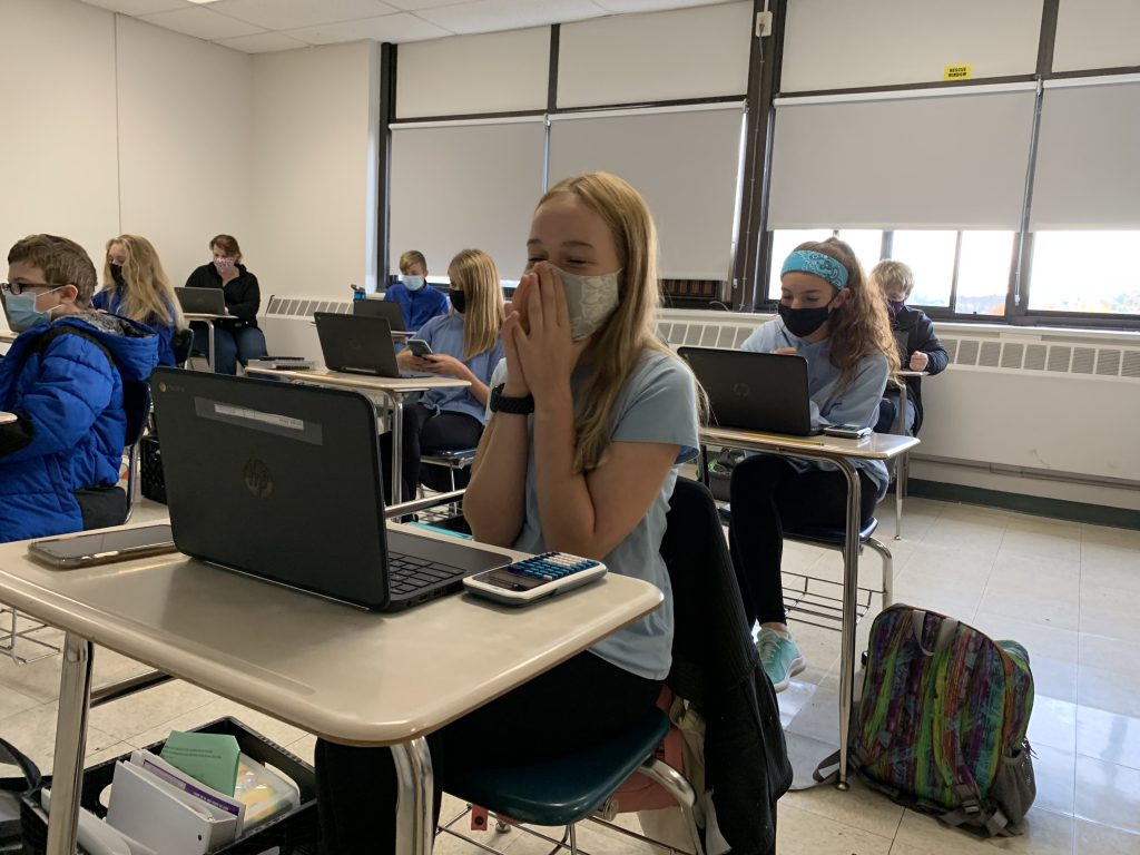 a student wearing a mask seated before a chromebook excitedly holds her hands up towards her face