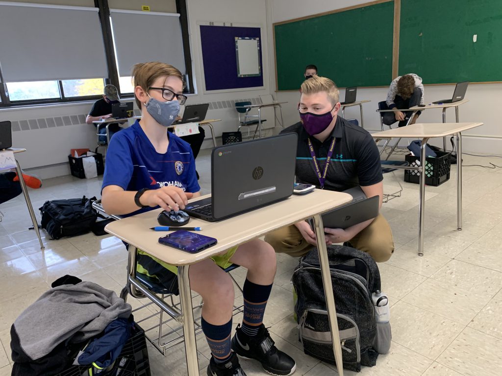 a teacher wearing a mask crouches next to a student seated at a desk before a chromebook