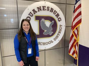 a woman wearing a blue shirt with a black vest stands in front of a logo for Duanesburg Central School, next to an American flag