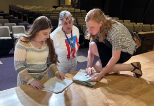 an adult woman and two teenage girls go through a play script on a theatrical stage