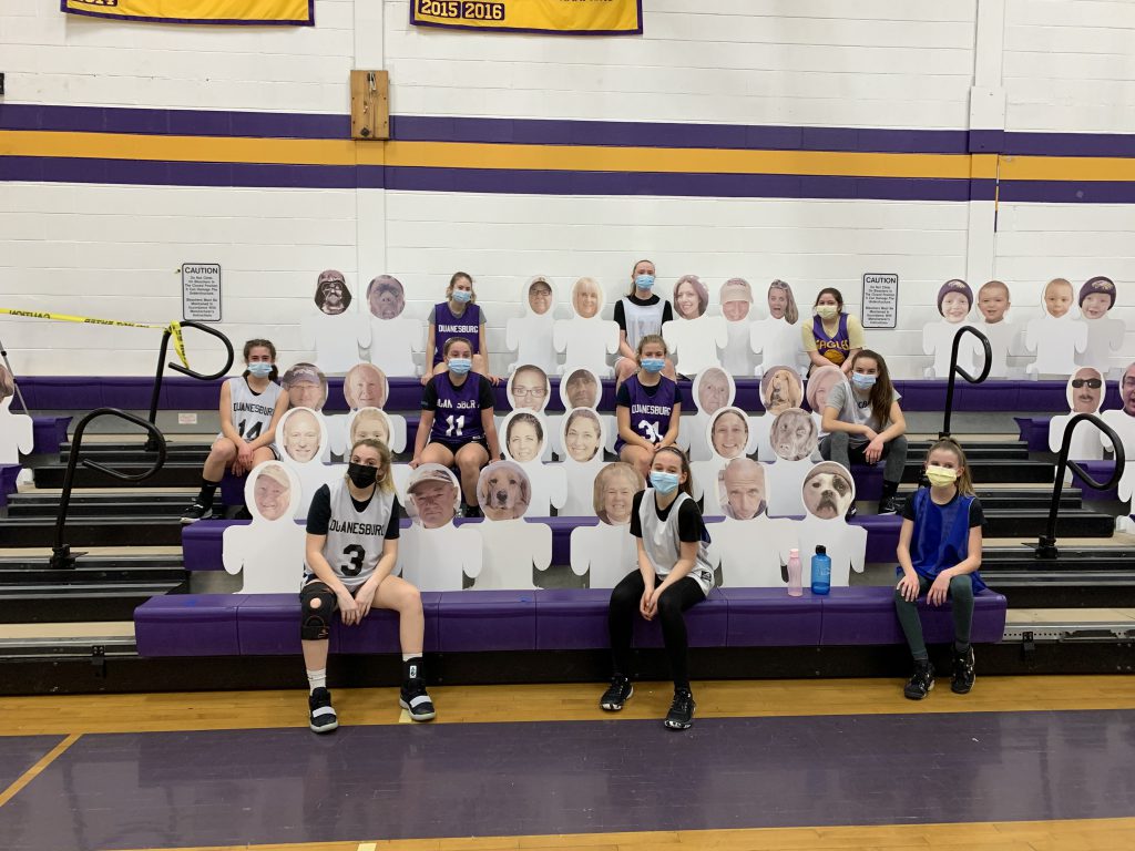 Girls junior varsity basketball teammates sit with cutouts in the stands of a school gymnasium