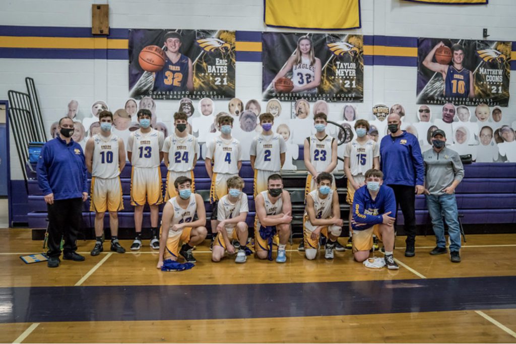 Boys varsity basketball teammates sit with cutouts in the stands of a school gymnasium