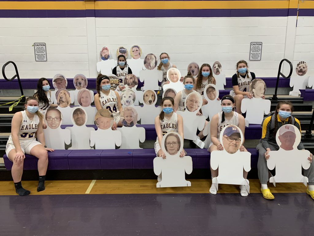 Girls varsity basketball teammates sit with cutouts in the stands of a school gymnasium