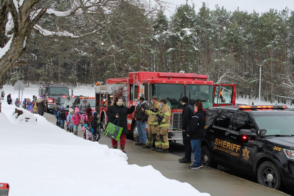 line of kindergarten students walk on a sidewalk in front of a row of firetrucks and police cars outside of a school