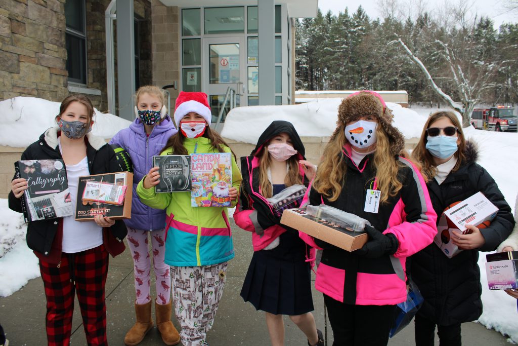 group of six students wearing masks and coats hold up gifts outside of an elementary school