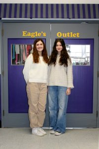 two teenaged girls with long dark hair stand outside of a storefront labeled "Eagle's Outlet."