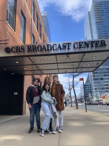 Four teenagers stand outside on a sidewalk, underneath a sign that reads CBS Broadcast Center