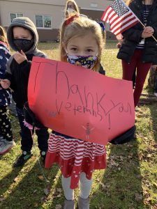 a young girl wearing red, white and blue holds a red handmade sign that says "Thank you veterans!"