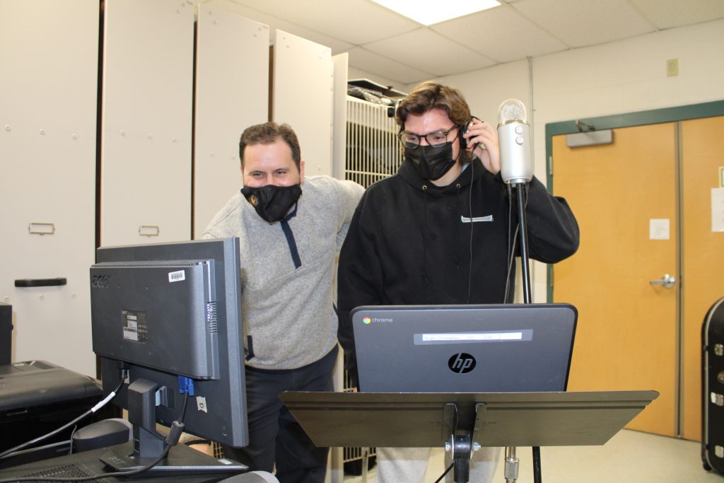 high school music teacher wearing a mask works on a computer while student wearing a mask holds headphones to his ear in a school classroom