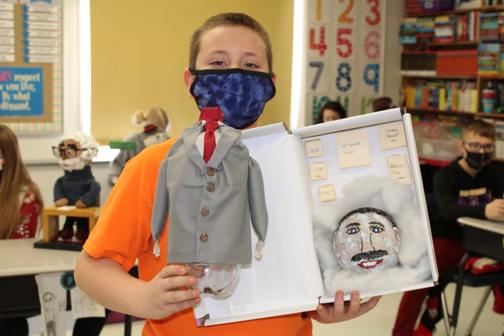 student with short hair wearing a mask holds a model made out of a soda bottle and a model freezer meant to portray a cryogenically frozen walt disney