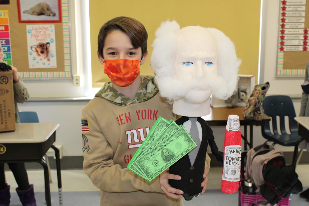 student with short hair and wearing a mask holds up a 3-d model made out of a soda bottle meant to depict mr. heinz