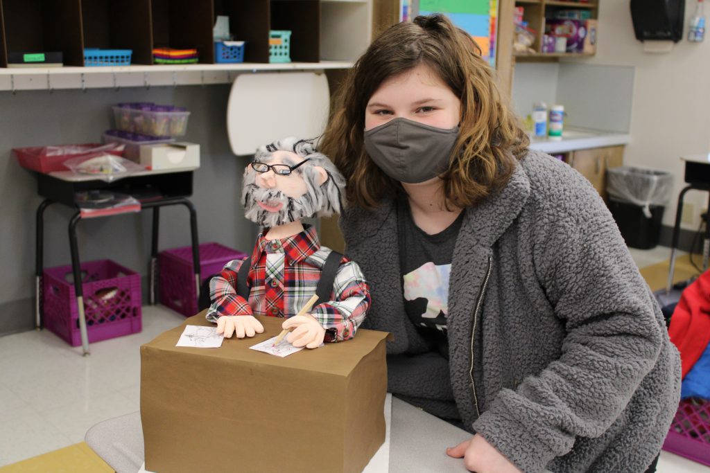student with shoulder length brown hair stands next to a 3d model meant to depict dr. seuss sitting at a writing desk