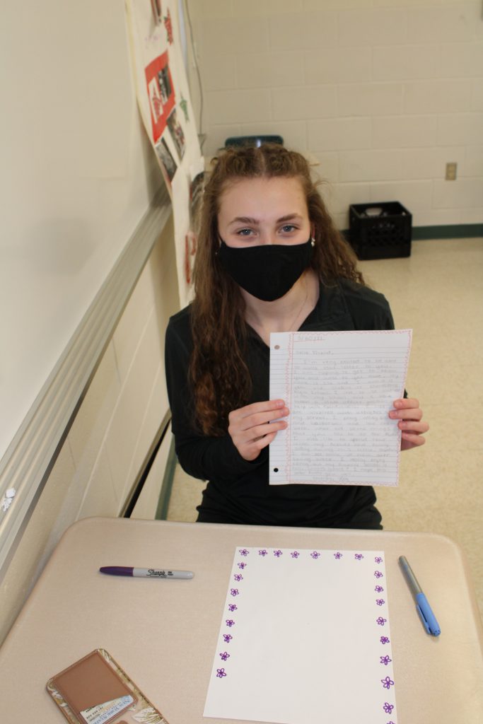 high school student with long hair wearing a face mask holds up a handwritten letter while seated in a desk in a classroom
