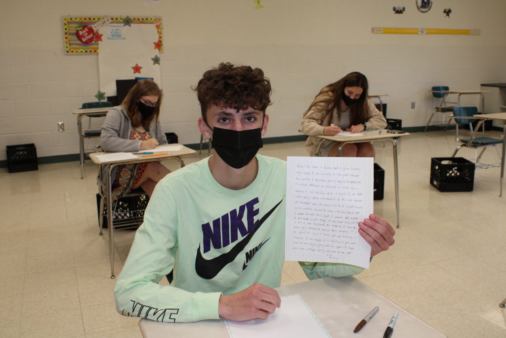 high school student with short curly hair wearing a face mask holds up a handwritten letter while seated in a desk in a classroom