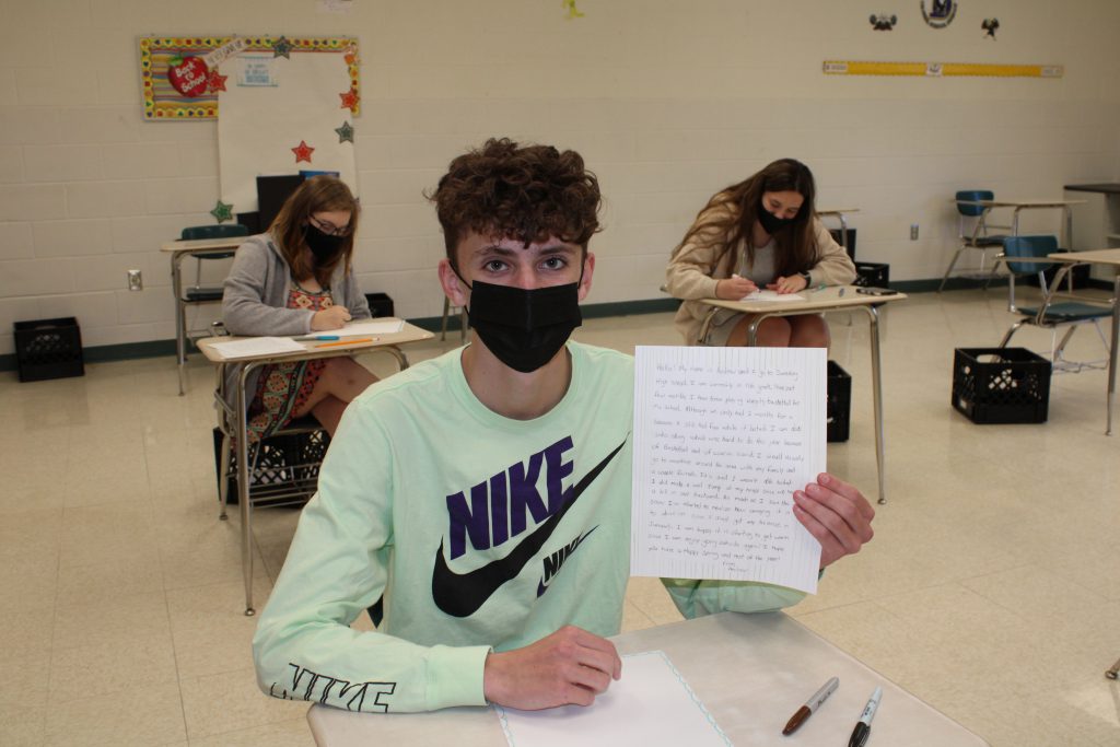 high school student with short curly hair wearing a face mask holds up a handwritten letter while seated in a desk in a classroom