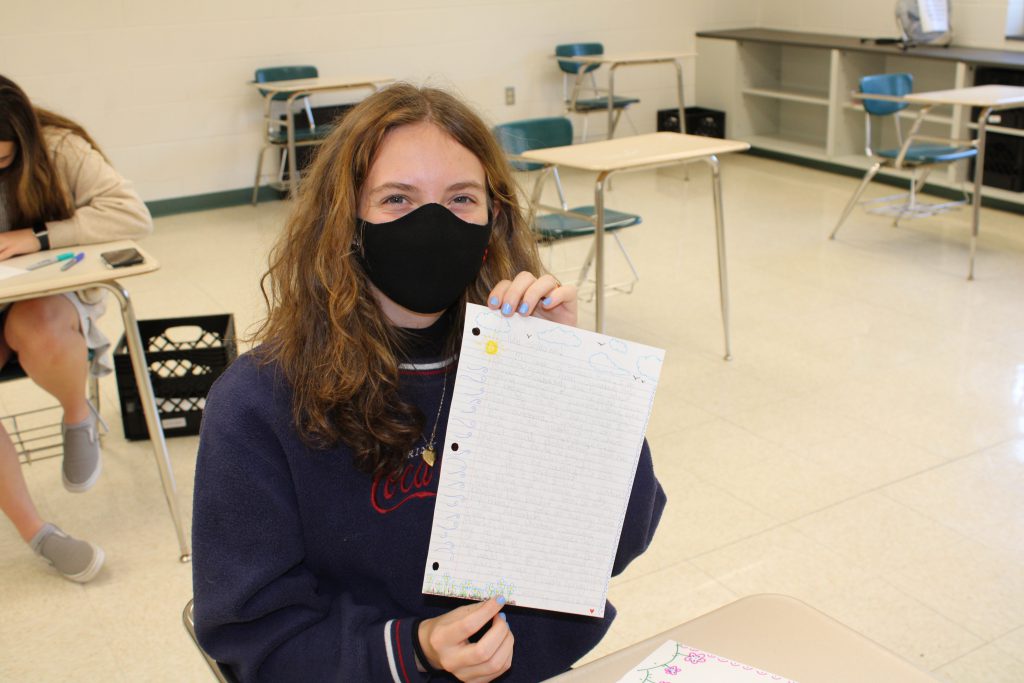 high school student with shoulder length curly hair wearing a face mask holds up a handwritten letter while seated in a desk in a classroom
