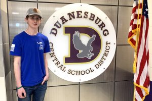 a young man wearing a baseball cap poses next to a Duanesburg School logo and the American flag