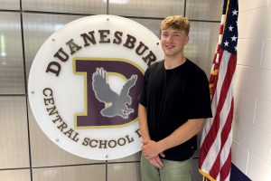 a tall young man in a black t-shirt stands in front of his school logo