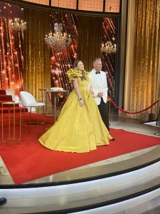 a woman wearing a yellow gown and a man wearing a white tuxedo pose on a stage with a red carpet