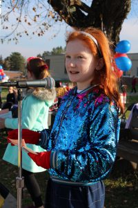 a young girl with red hair and a sparkly blue coat speaks into a microphone