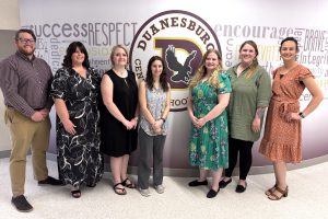 seven men and women pose in a hallway, in front of a school logo