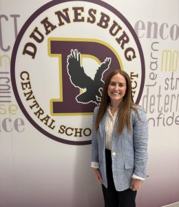 a young woman wearing a light blue blazer poses in front of a school logo