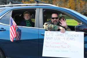 a male veteran wearing sunglasses and holding a sign, waves to students from his vehicle