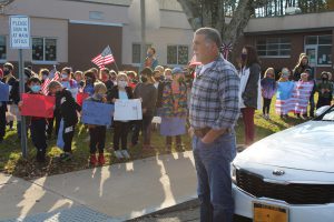 a veteran wearing blue jeans and a plaid shirt watches the Veterans Day ceremony from a distance, with elementary students in the background