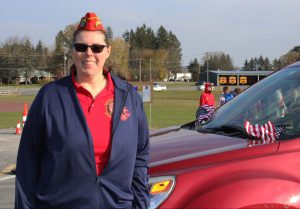 a female veteran wearing a red shirt, blue coat, red hat and sunglasses stands next to a red car