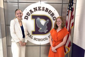 two young women stand in front of a school logo and the American flag