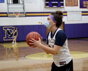 a girl wearing a face mask, purple headband and a white jersey throws a basketball
