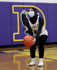 a young female athlete wearing a face mask and white basketball jersey throws a basketball