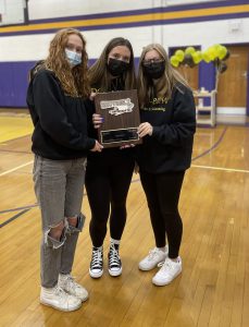 three female students wearing face mass pose inside a gymnasium holing a plaque
