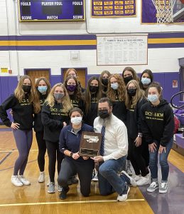 a group of female students wearing face masks pose with two adults holding a plaque, inside of a gymnasium
