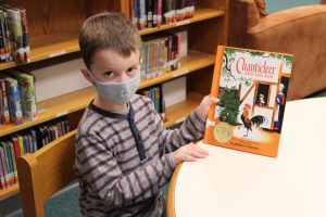 a boy wearing a face mask shows his book while sitting at a table in the library