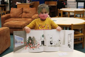 a girl wearing a face mask shows off her book while in the library