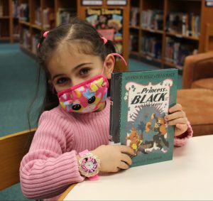 a young girls wearing a face mask shows her book while in the library