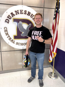 a man wearing a shirt that says "Free Hugs" stands in front of a school logo