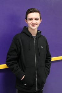 a young man wearing a black hoodie stands in front of a purple wall.