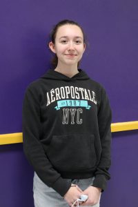 a young woman wearing a black sweatshirt stands in front of a purple wall.